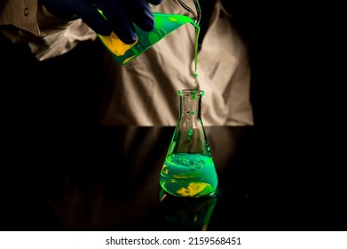 A Woman Scientist Experimenting With A Green Fluorescent Solution In A Glass Conical Flask In Dark Biomedical Laboratory For Health Care Medicine Development. Copy Space Black Background