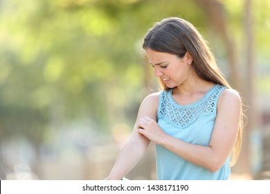 Woman scratching arm because it stings in a park with a green background