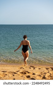 Woman sea swimsuit sand. A girl in full growth stands with her back and enters the sea in a black swimsuit. Alone on the beach on a sunny day. - Shutterstock ID 2248492495