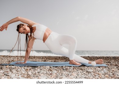 Woman sea yoga. Well looking middle aged woman with braids dreadlocks in white leggings and tops doing stretching pilates on yoga mat near sea. Female fitness yoga routine concept. Healthy lifestyle. - Shutterstock ID 2255554499