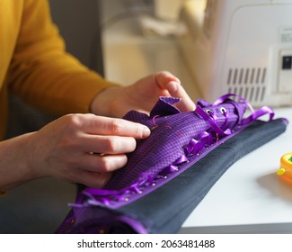 Woman Seamstress Or Tailor Using Sewing Hook While Making Purple Corset With Lacing, Working At Her Creative Workplace In Atelier Or Home With Colored Pins On Table. Lingerie Manufacture Concept