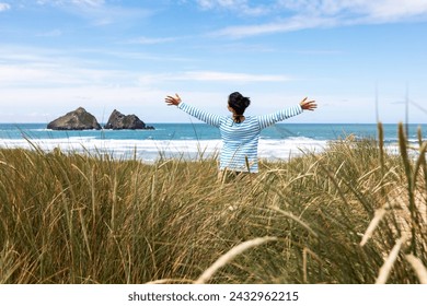 Woman at seaside in Cornwall looking at sea and raising arms - Rear view of a woman standing on sand dunes with ocean on background on a sunny day - Travel and happiness concepts - Powered by Shutterstock