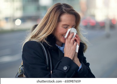 Woman with a seasonal winter cold blowing her nose on a handkerchief or tissue as she walks down an urban street in a health and medical concept - Shutterstock ID 650987659
