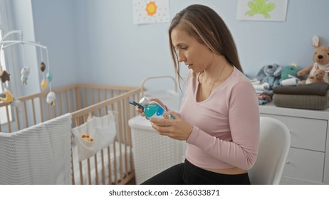 Woman seated in cozy bedroom examining baby bottle near cradle in brightly lit home interior, with soft toys scattered, embodying serene motherhood moment.