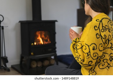 Woman Seated On The Floor Enjoying Her Hot Drink In Front Of A Wood Burning Stove Or Fireplace.