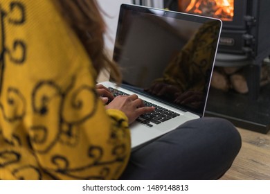 Woman Seated On The Floor Working On Her Laptop Computer In Front Of A Wood Burning Stove Or Fireplace.