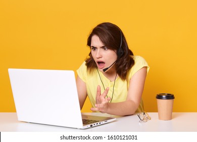 Woman seatting at table and looking at her laptop screen in consternation and horror, keeping mouth opened, wearinfcasual tshirt, posing isolated over yellow studio background, working online. - Shutterstock ID 1610121031