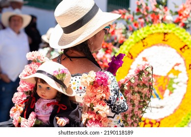 Woman Seen From The Back With Hat And Typical Costume Of Colombian Culture At The Flower Festival. Person Carries A Doll On Her Back In A Colorful Parade Full Of Flowers.