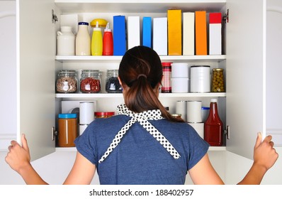 Una mujer vista desde atrás abriendo las puertas a una despensa llena. El armario está lleno de varios alimentos y comestibles, todos con etiquetas en blanco. El formato horizontal de la mujer es irreconocible.