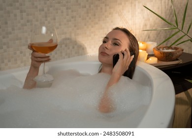 A woman is seen enjoying a relaxing evening in the bathtub filled with bubbles. She is holding a glass of wine and talking on the phone. The warm glow of candlelight creates a cozy atmosphere - Powered by Shutterstock