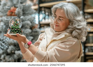 Woman selecting Christmas gifts in store, holding in front of her a snow globe figurine with a Christmas tree with snow inside it - Powered by Shutterstock