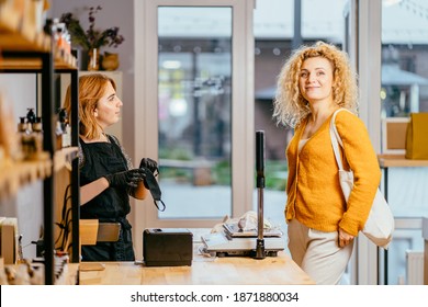 Woman Seller At Counter Checkout Of Modern Sustainable Plastic Free Grocery Store. Female Customer With Shopping Reusable Cotton Bags. Seller And Buyer On Grocery Store Interior.