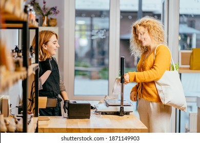 Woman Seller At Counter Checkout Of Zero Waste Shop Female Customer With Shopping Reusable Cotton Bags. Girl With Purchases At Cashier Desk. Seller And Buyer On Grocery Store Interior.