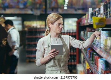 A woman seller in a supermarket uses a tablet computer to count the remaining goods, colleagues conduct an inventory in the grocery department among the shelves with goods - Powered by Shutterstock