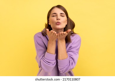 Woman sending air kiss to camera, flirting and demonstrating love affection feelings, keeps eyes closed, romantic relationships, wearing purple hoodie. Indoor studio shot isolated on yellow background