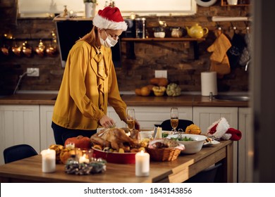 Woman Serving For At Dining Table For Christmas Meal And Wearing Protective Face Mask Due To Coronavirus Pandemic. 