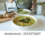 Woman serving with a ladle a delicious homemade, vegetarian pea soup with mushrooms in the kitchen