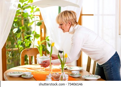 Woman Setting The Table For Tea Of Coffee Time In Her Domestic Dining Room, The Table Is Decorated With Tulip Flowers, Whole Scene Is Sunlit