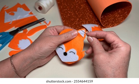 Woman sews toy fox ear to its muzzle, creating felt toy. Hands of craftswoman at home sew up fox toys made of orange felt. Hobby of creating toys for children - Powered by Shutterstock