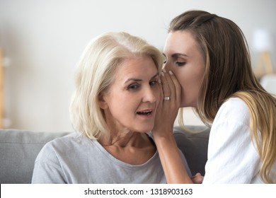 Woman Sharing Secret With Surprised Aged Mother, Daughter Whispering In Old Mum Ear, Hearing Shocking News, Family Gossiping At Home, Sitting Together On Couch, Best Friends Close Up