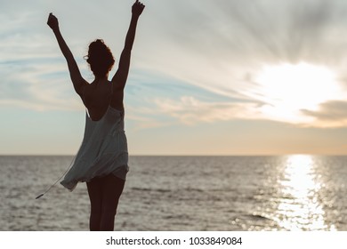 woman in sheer dress at sunset facing ocean celebratory pose - Powered by Shutterstock