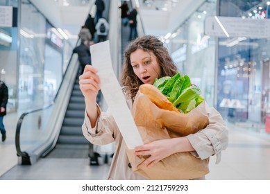 A woman in shock looks into a paper check from a supermarket in a shopping center against the background of an escalator and holds a package with fresh products, price increase - Shutterstock ID 2121759293