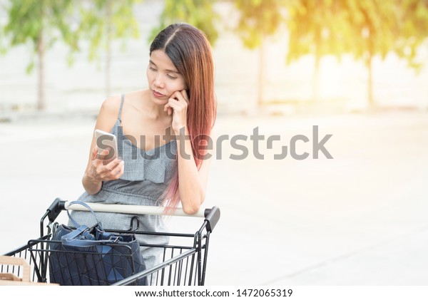 Woman with shopping cart at car\
park and use smart phone with thinking position,business asian\
woman look at smart mobile phone with depresses and\
headache.