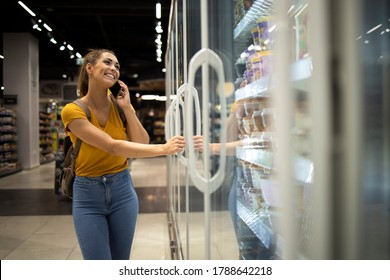 Woman With Shopping Cart Opening Fridge To Take Food In Grocery Store While Talking On The Phone. Woman Buying Groceries In Supermarket.