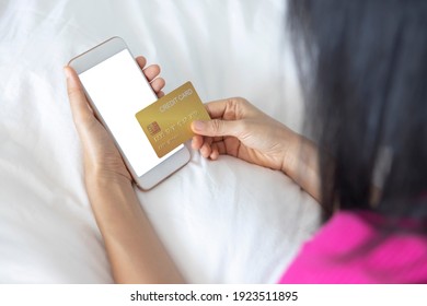 A Woman Shopping Online On Her Bed At Home With A Smartphone Using A Gold Credit Card. And Use Your Credit Card To Tap The Screen To Verify The Identity. Concept About Business. Over Shoulder Shot.