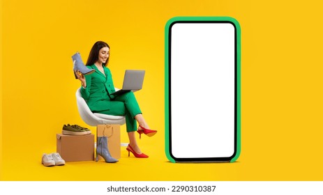 Woman shopping online via laptop, sitting near huge phone with blank screen, holding new shoes over yellow studio background. E-commerce, fashion and technology. Mockup - Powered by Shutterstock