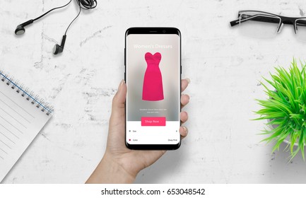 Woman Shopping With Smart Phone. Pink Woman Dress On Ecommerce App. White Desk In Background. Plan, Glasses, Headset Beside.