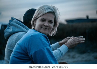 A woman with short blonde hair and a blue jacket smiles at the camera while sitting next to a man in a gray hoodie. They are outside by the Baltic Sea at dusk with a blurred background of fields and b - Powered by Shutterstock