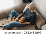 A woman with short blonde hair relaxes on a sofa with her dog, enjoying a peaceful moment at home. The cozy setting and loving companionship create a feeling of comfort and happiness.