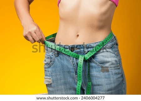 Woman with short hair in oversized jeans and measure tape isolated on yellow background. Weight loss and healthcare concept