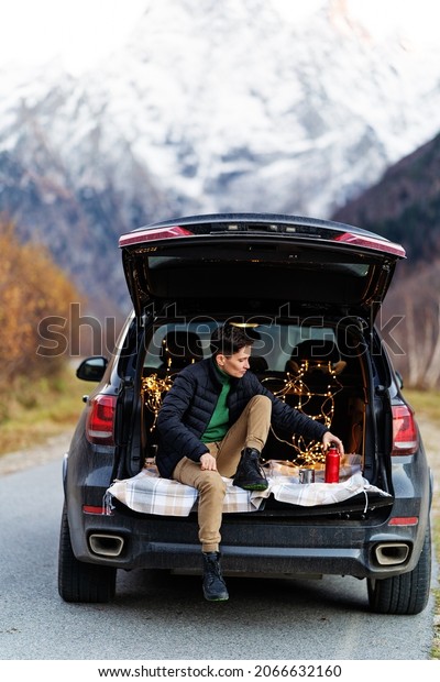 A woman with a short\
haircut in a sweater sits in the trunk of a car against the\
background of mountains. Garland lights on the background. High\
quality photo