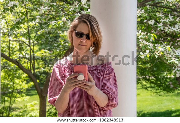 Woman with short streaked hair and off shoulder\
blouse taking a selfie of herself in pagoda surrounded by dogwood\
blossoms and spring\
foliage