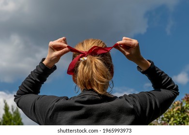 A woman shot from behind makes a red bow to tie her blonde hair in which there is also a daisy flower, against the sky