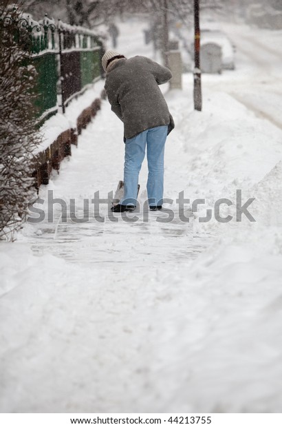 Woman shoveling snow from the\
sidewalk in front of her house after a heavy snowfall in a\
city