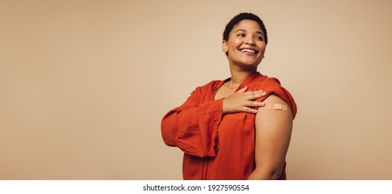 Woman Showing Her Arm After Receiving Vaccine Shot On Brown Background. Female With Bandage On Her Arm Looking Away And Smiling.