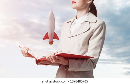 Woman Showing Rocket Ship On Open Book. Rocket Launch As Symbol Startup Company. New Creative Project Concept. Woman In White Business Suit On Background Of Skyscape. Business Innovation.