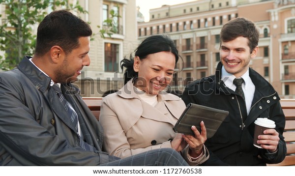 The\
woman is showing something on the tablet. The whole company is\
discussing this with interest. Two men and a woman are sitting on a\
bench. A woman is holding a tablet in her\
hands.