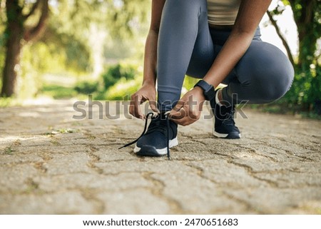 A woman is shown tying her shoelaces while standing on a path. She is focused on the task at hand, ensuring her shoes are securely fastened before continuing her walk, cropped