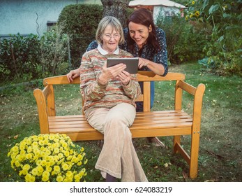 Woman Shows Her Mother Something On The Tablet. They Are Sitting Outside In The Garden.