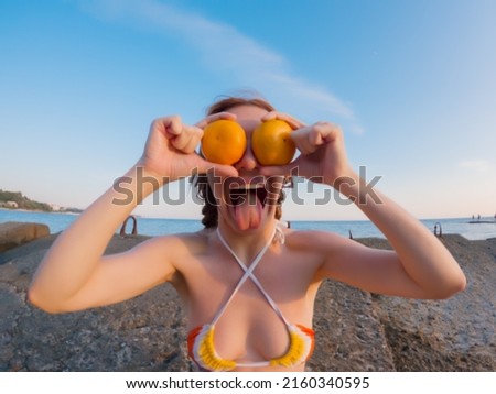 A woman shows her tongue putting tangerines to her eyes sitting on the breakwaters of the sea coast on a summer day. Humorous photo. Wide angle view through go pro camera. Unfocused view.