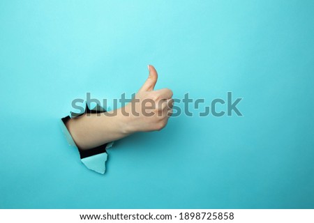 Woman shows like gesture through torn blue wall, keeps thumb up, says you are best, demonstrates approval sign, recommends something. Copy space aside for your advertising content