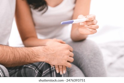 A woman shows a pregnancy test to her partner in bed, with a positive result indicated. The couple is sitting up in bed, and the woman is holding the test up in front of her partner, cropped - Powered by Shutterstock