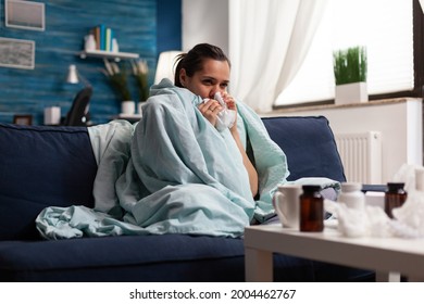 Woman With Sickness Wrapped In Blanket At Home Ill Feeling Unwell. Young Caucasian Adult Suffering From Temperature Fever Disease Illness Flu, Taking Medical Treatment Sitting On Couch