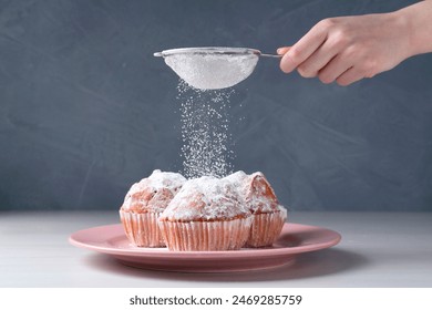 Woman with sieve sprinkling powdered sugar onto muffins at white wooden table, closeup