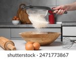 Woman sieving flour into bowl at white wooden table in kitchen, closeup