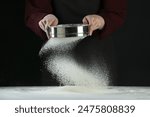 Woman sieving flour at table against black background, closeup